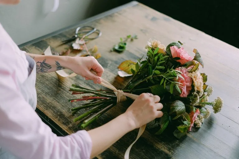 A person's hands are shown tying a bouquet of flowers with a ribbon on a wooden table. The bouquet contains a variety of flowers including pink roses, and the table has scissors and additional cut flowers scattered around, suggesting a DIY floral arrangement process.